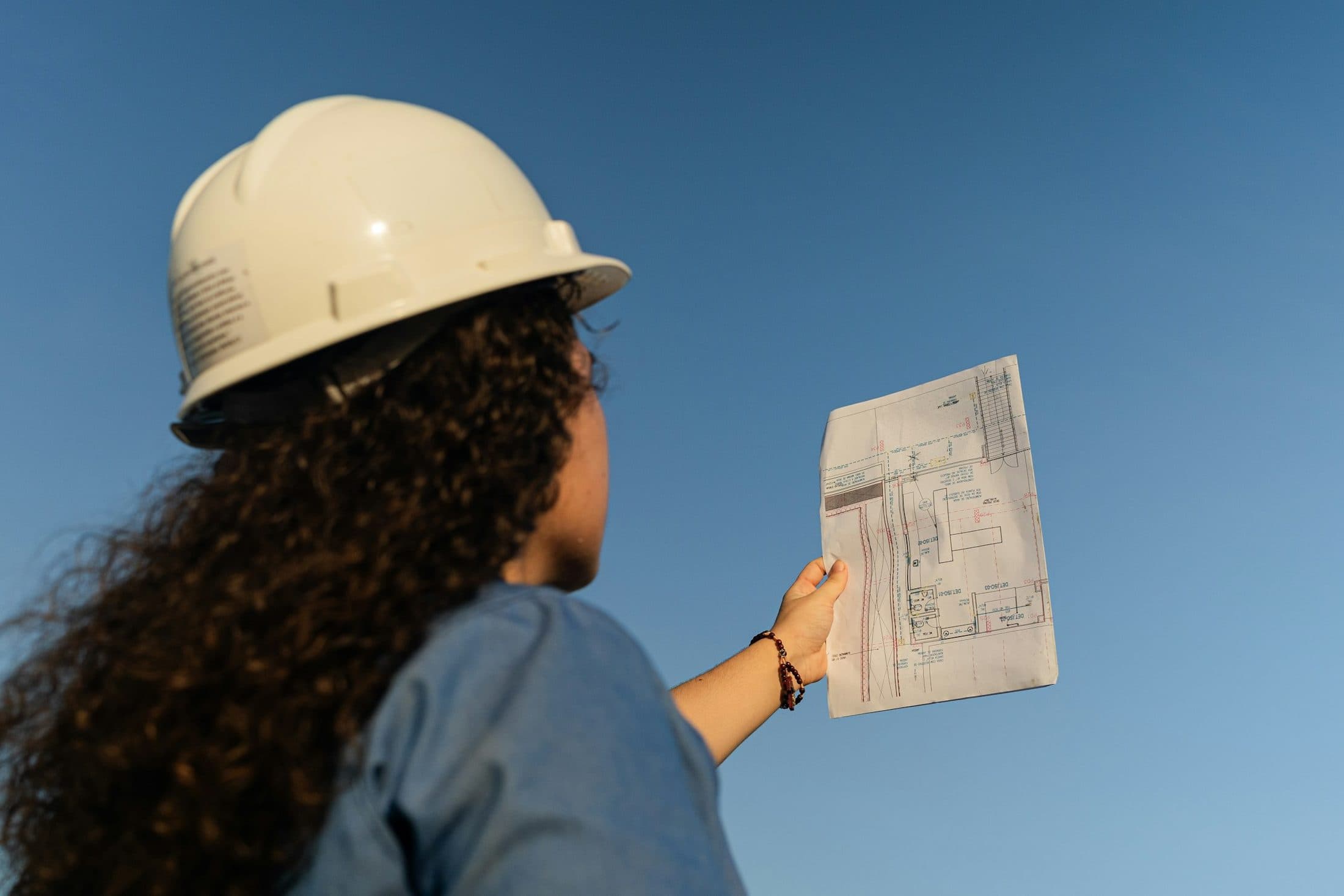 Une conductrice de travaux vue de dos, casque sur la tête, un plan de chantier levé devant elle
